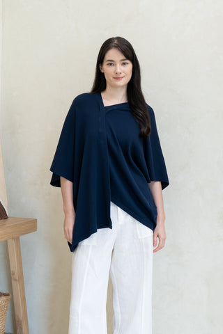 Indoors, a woman wears Olive Basics’ Deborah Nursing Kimono Cardigan in navy blue and white wide-leg pants. Barefoot by neutral decor and a basket, she uses the kimono as both a stylish layer and breastfeeding cover.