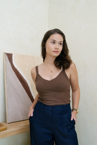 A woman wearing the Olive Basics Diana V-neck Reversible Padded Tank in brown and dark blue pants stands indoors with her hands in her pockets, looking to the side.