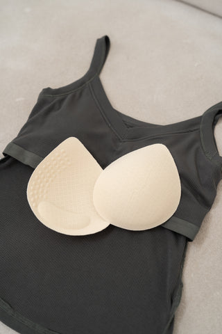 A pair of beige bra inserts sits atop the Olive Basics Diana V-neck Reversible Padded Tank in dark gray ribbed modal knit, laid flat on a light surface.