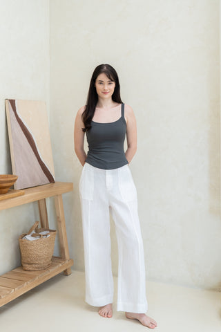 A woman with long dark hair, barefoot in a minimalist room, wears the Olive Basics Diana V-neck Reversible Padded Tank and white pants, standing beside a wooden table decorated with ornaments.