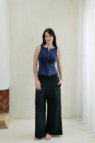A woman stands indoors by a window with white curtains, wearing the Olive Basics Grace Reversible Button Tank in navy and wide-leg black pants, with one hand in her pocket.