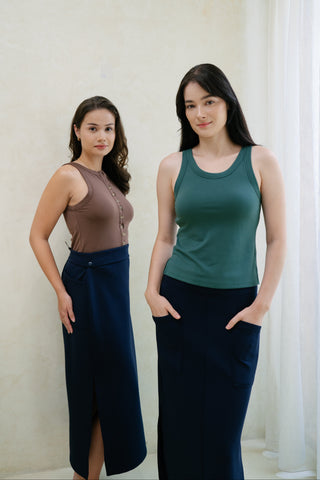 Two women stand indoors near a white curtain, both wearing Olive Basics Grace Reversible Button Tanks and navy blue skirts, facing the camera with neutral expressions.