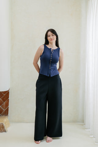 Woman standing indoors by a window, barefoot with hands in pockets, wearing Olive Basics Isabel Expandable Hi-Low Pocket Pants in black and a sleeveless navy top.