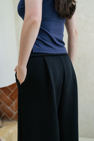 Model wears Olive Basics Isabel Expandable Hi-Low Pocket Pants in black with an adjustable waistband and pockets, styled with a navy sleeveless top, standing with hands in pockets and facing slightly away from the camera.