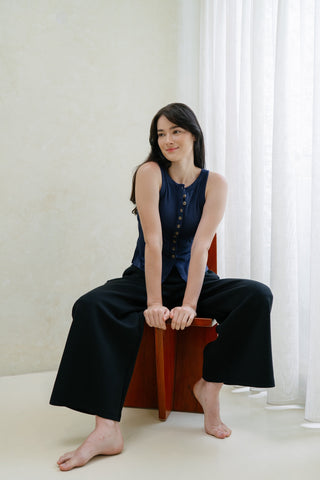 A woman with long dark hair sits barefoot on a wooden chair, wearing Olive Basics' Isabel Expandable Hi-Low Pocket Pants in black and a navy sleeveless top, in a light room with sheer curtains.
