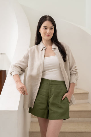 A woman with long dark hair stands on a staircase, wearing the Olive Basics Marie Everyday Shirt in TENCEL™ Linen over a white top and olive green shorts—one hand on the railing, the other in her pocket for an effortless look.
