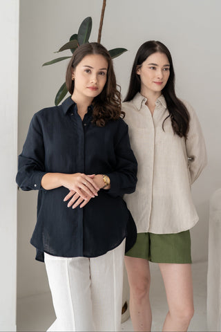 Two women stand indoors near a plant; one wears a tailored black shirt and white pants, while the other showcases casual style in the Olive Basics Marie Everyday Shirt in TENCEL™ Linen paired with green shorts.