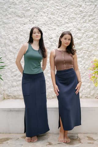 Two women stand barefoot outdoors against a textured white wall, wearing sleeveless tops and Olive Basics Michal Reversible Maxi Skirts with adjustable waistbands. Shrubs frame the scene.