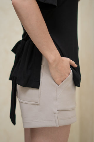 Model wearing Olive Basics Naomi Stretchy Pocket Shorts in light grey, paired with a black top and one hand in the pocket, shown from the waist down against a neutral background.