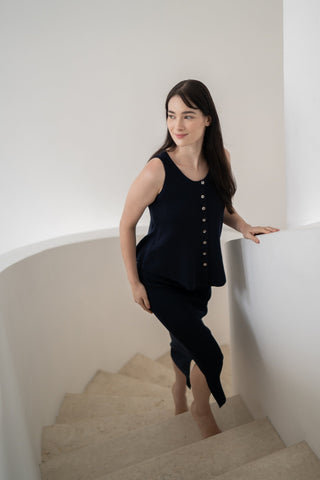 Wearing the Leah Super Stretch Slit Skirt by Olive Basics, a woman in a black sleeveless top walks up a curved white staircase, hand on the railing and looking to the side.