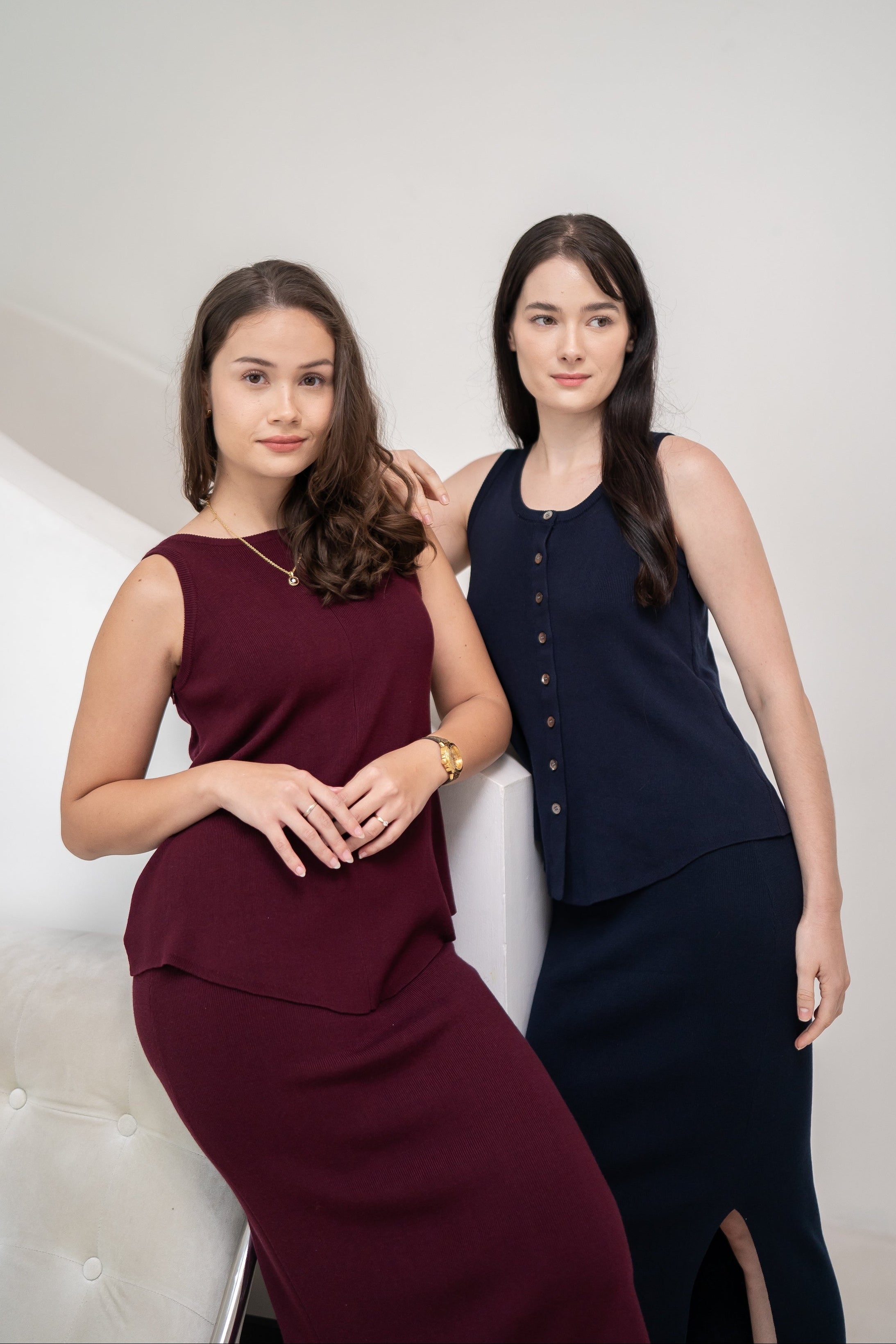 Two women with long dark hair pose indoors against a white background; one wears a burgundy sleeveless dress, while the other showcases an Olive Basics Leah Super Stretch Slit Skirt paired with a navy sleeveless top for effortless comfort.