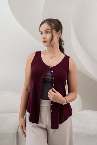 A woman stands indoors before a modern white backdrop, wearing the Olive Basics Sarah Reversible Button Tank in burgundy with light beige pants.