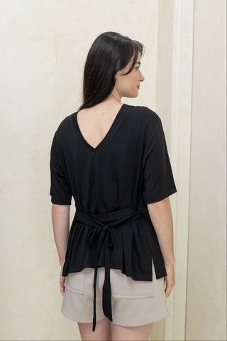 A woman with long dark hair wears the Olive Basics Tabitha Reversible Multi-way Tee in black and beige adjustable-waist shorts, standing indoors by a cream wall and door with shelves in the background.