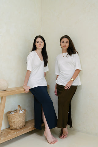 Two women in Olive Basics' Tabitha Reversible Multi-way Tees and long skirts with adjustable waists stand barefoot indoors near a light wooden bench with a basket and decorative orb.