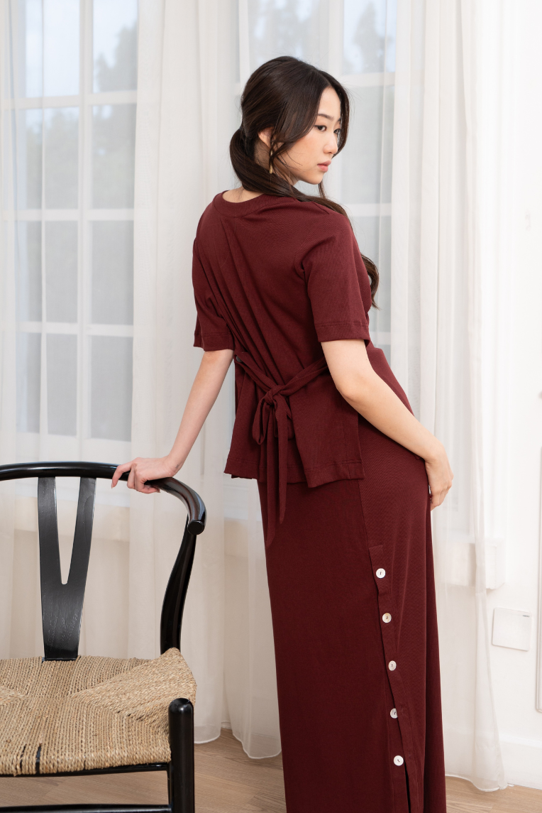 A woman models the Olive Basics Tabitha Stretchy Button Maxi Skirt, featuring an adjustable elastic waistband, as she stands beside a black chair in a bright room with sheer white curtains.