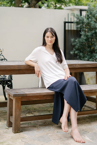 A woman with long dark hair sits barefoot on a wooden bench outdoors, wearing the Olive Basics Tabitha Reversible Multi-way Tee in a silky Modal blend and a dark skirt, with greenery and a white wall behind her.